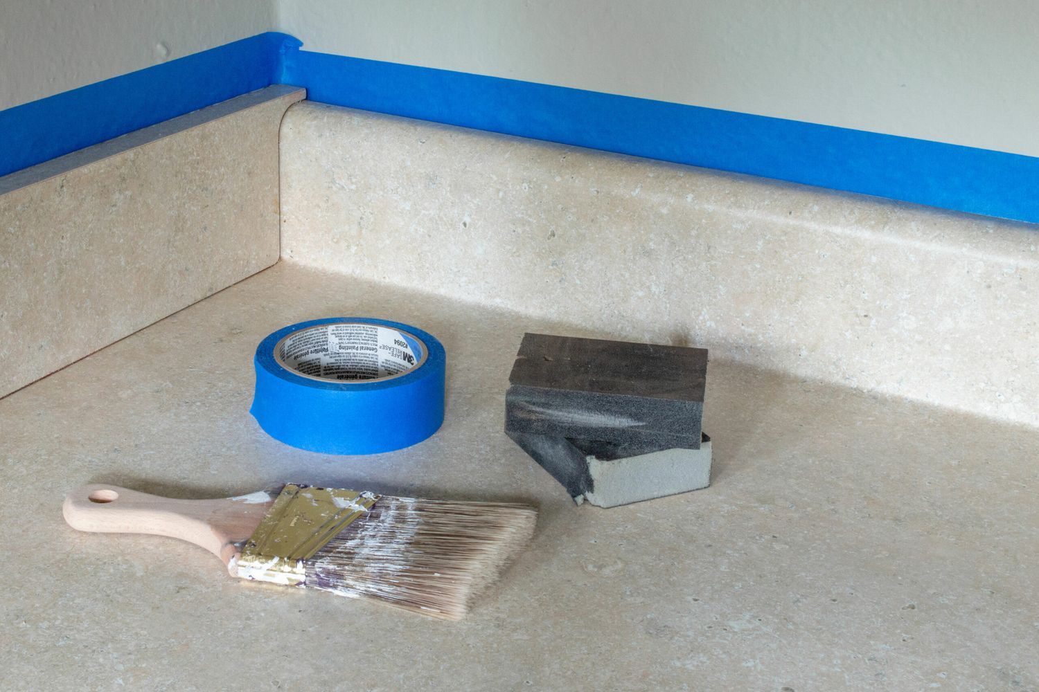 The supplies needed to prepare a laminate countertop, including a paint brush and blue tape.