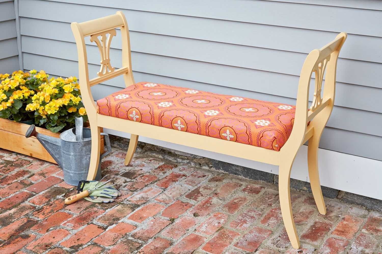 An outdoor bench with a pink cushion sitting next to a watering can and flowers.