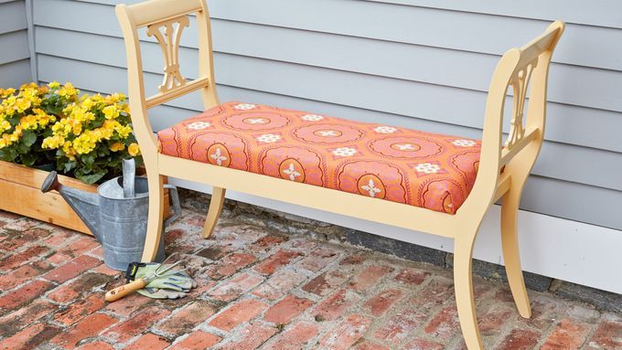 An outdoor bench with a pink cushion sitting next to a watering can and flowers.