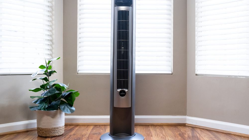 Lasko oscillating tower fan in front of a bay window, tan walls, and standing on a hardwood floor. Lead image for the best tower fan buying guide.