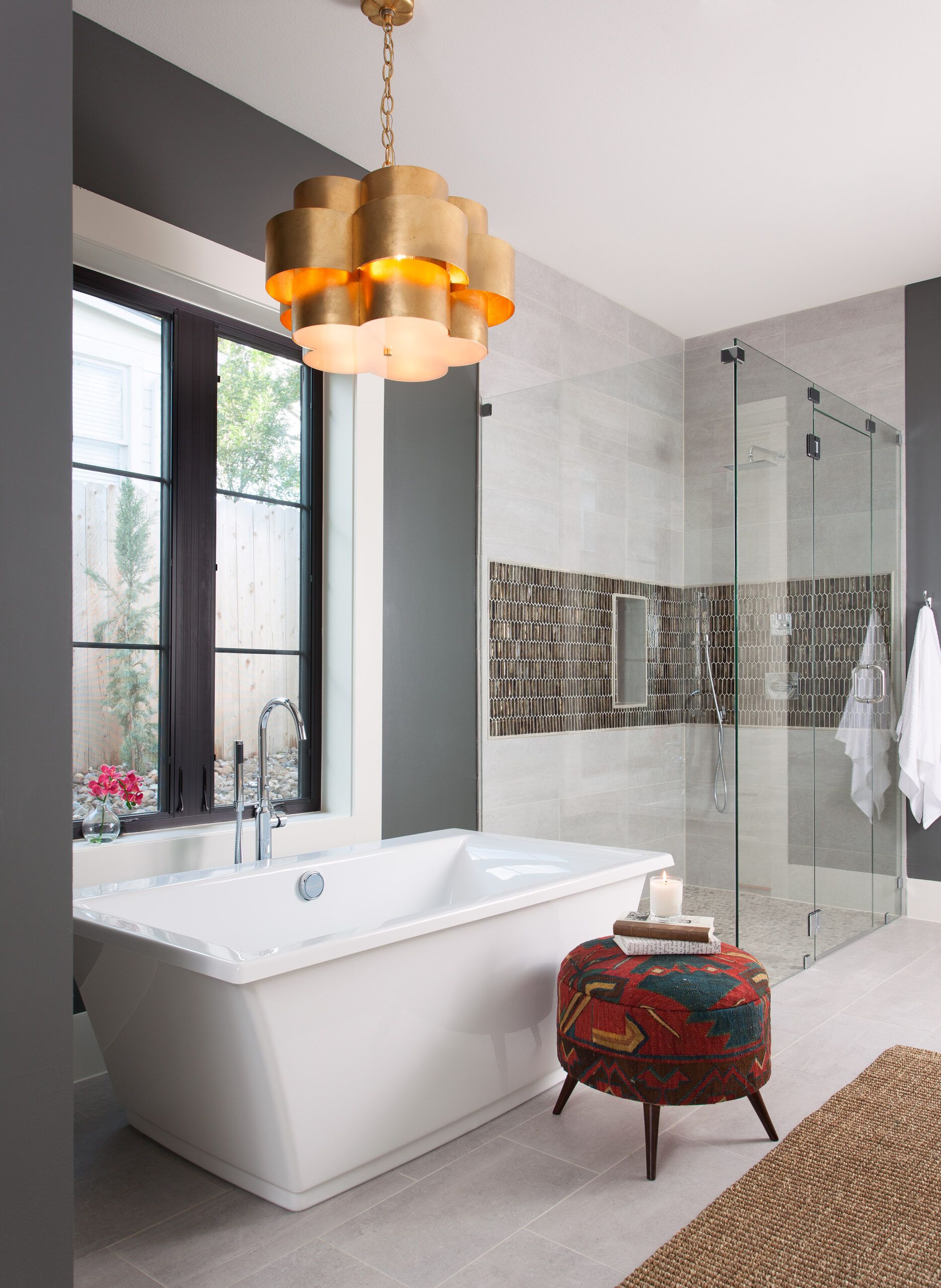 A modern bathroom featuring a unique gold-colored bathroom chandelier, a freestanding bathtub, a glass-enclosed shower, and a patterned ottoman.