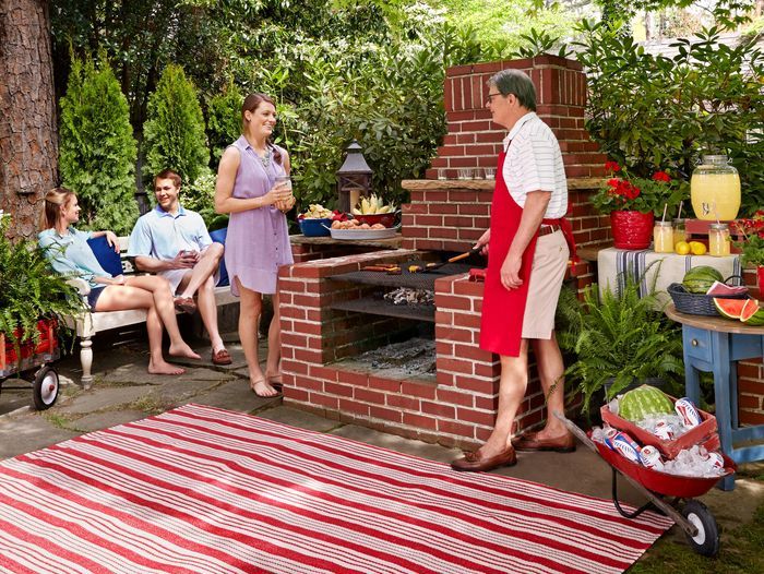 A group of people sitting around a brick barbecue pit. A man in a red apron is using it to cook.