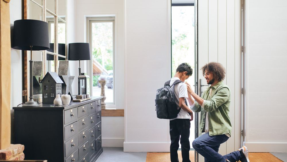 Lifestyle image inside the front entrance of a home showing a parent kneeling to help a young child with their backpack as they prepare to head back to school.