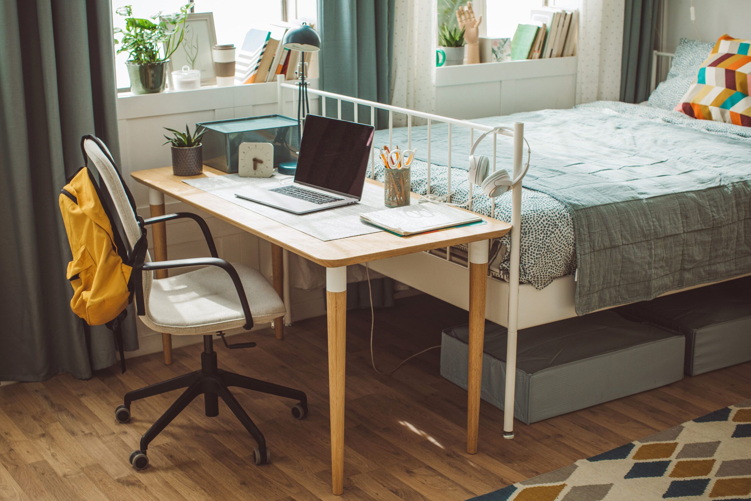 Lifestyle image of a dorm rooms with a bed, wooden writing desk, yellow backpack, chair, and laptop