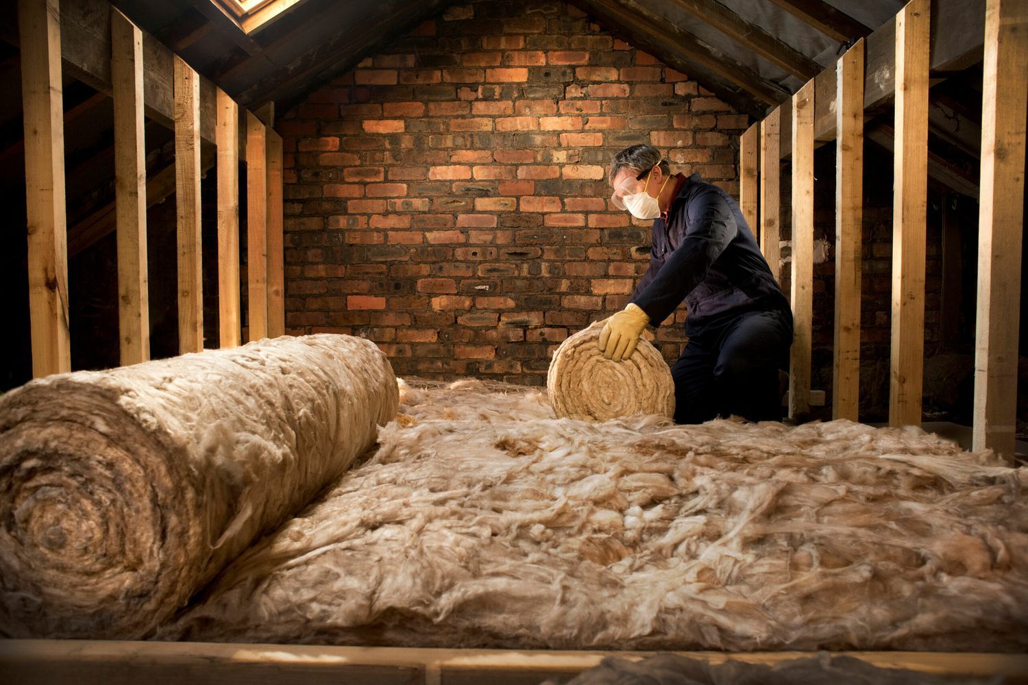 A worker in a protective mask and gloves installing rolls of fiberglass insulation, one of the common types of insulation, in an attic space.