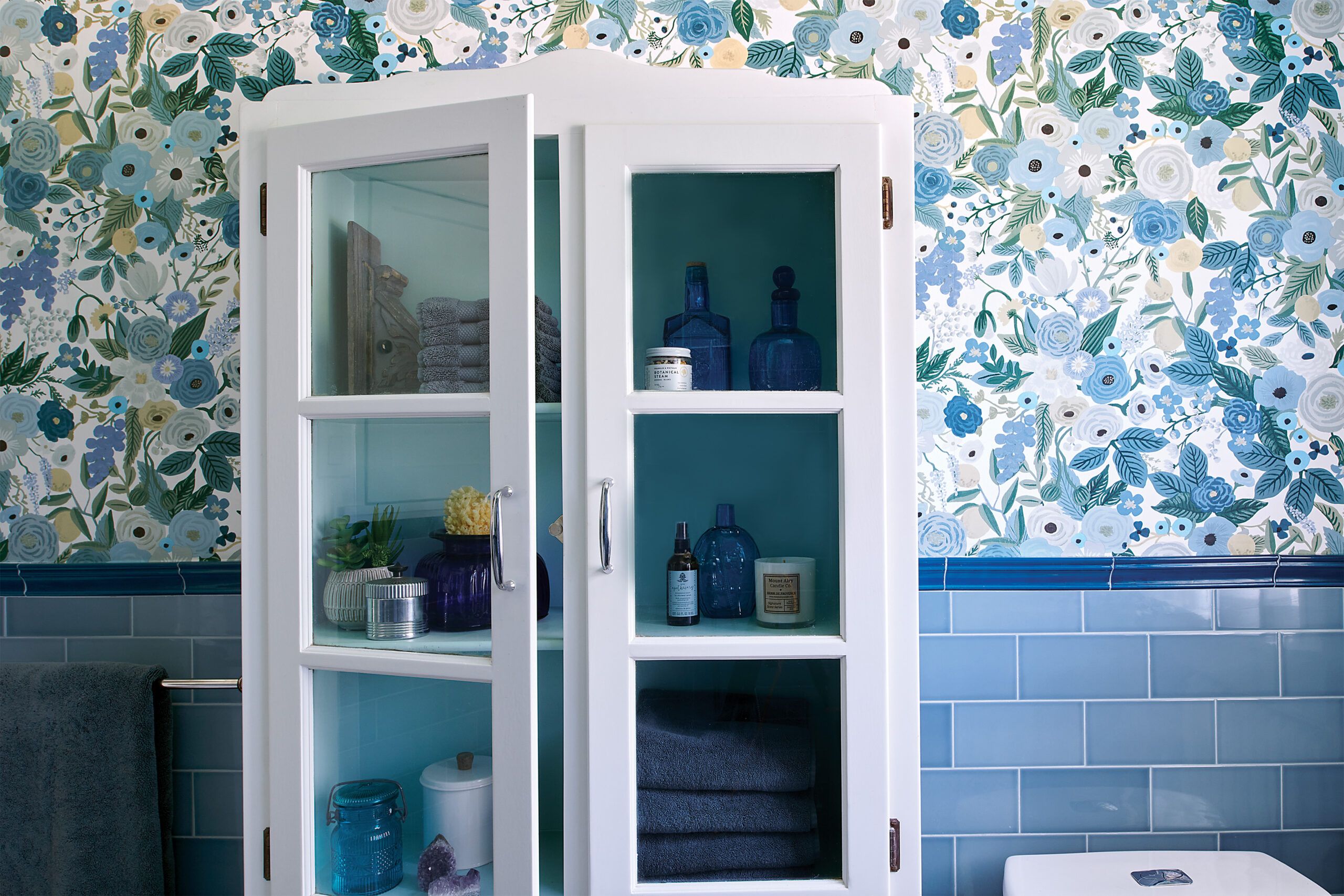 A vintage-inspired bathroom design, featuring a cabinet with glass doors that holds various items like towels, blue glass bottles, and toiletries. The wall behind it features a vibrant floral wallpaper in shades of blue, white, and green. The lower half of the wall is covered with blue tiles.