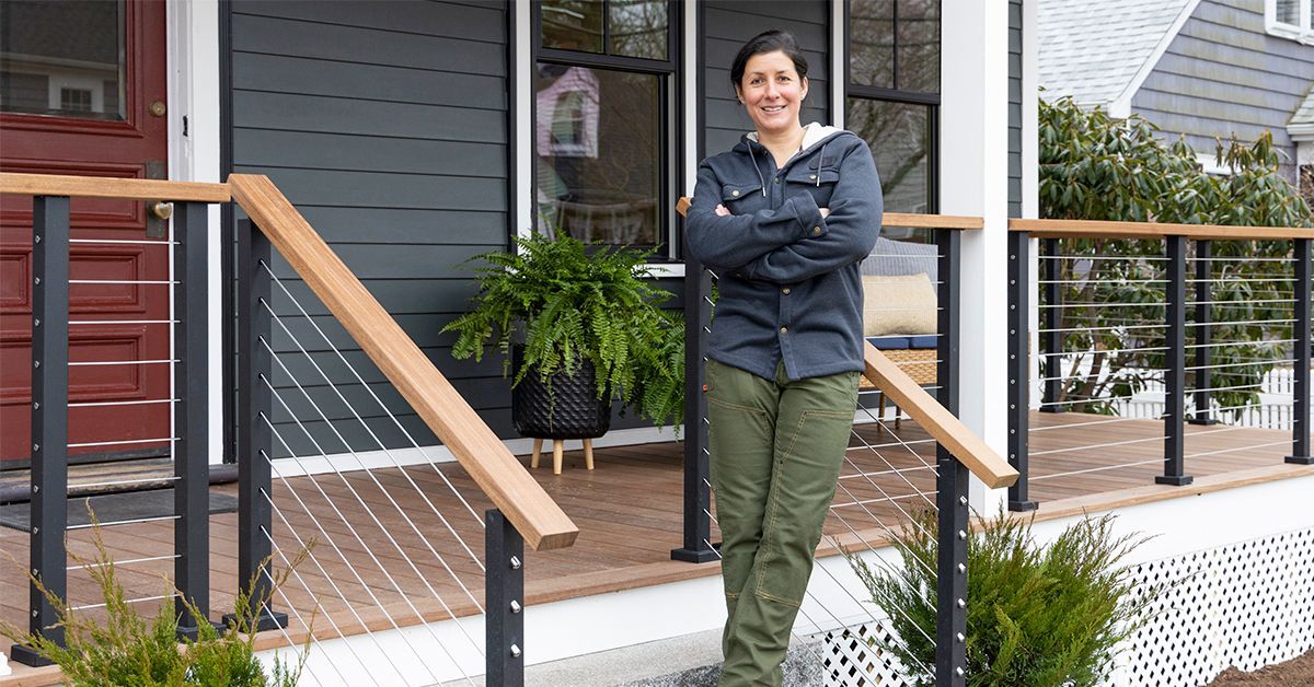 Landscape Contractor Jenn Nawada stands on the steps of a porch, arms crossed and smiling. They are wearing a dark jacket, green pants, and brown boots. The porch has cable railing with wooden handrails and potted plants beside the steps.