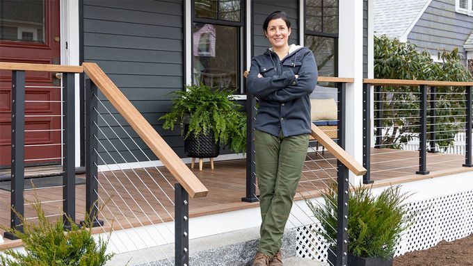 Landscape Contractor Jenn Nawada stands on the steps of a porch, arms crossed and smiling. They are wearing a dark jacket, green pants, and brown boots. The porch has cable railing with wooden handrails and potted plants beside the steps.