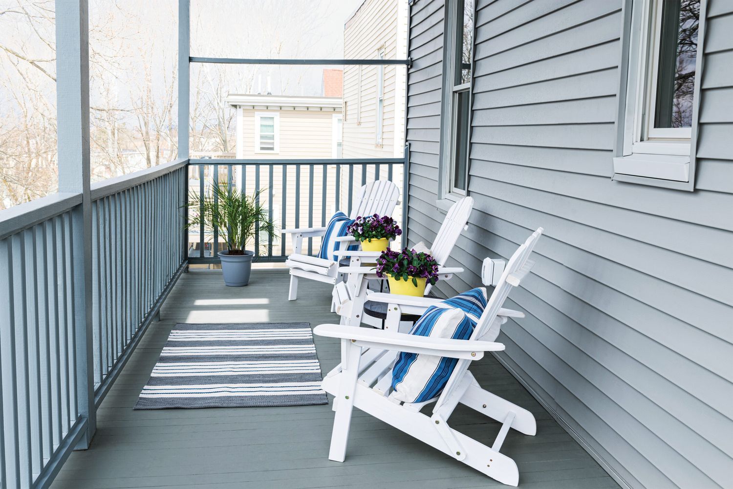 A cozy porch with two white Adirondack chairs, blue striped cushions, and a small table showcasing home maintenance tips to tackle this summer to keep everything inviting. A striped rug adds charm while a green plant sits in the corner.