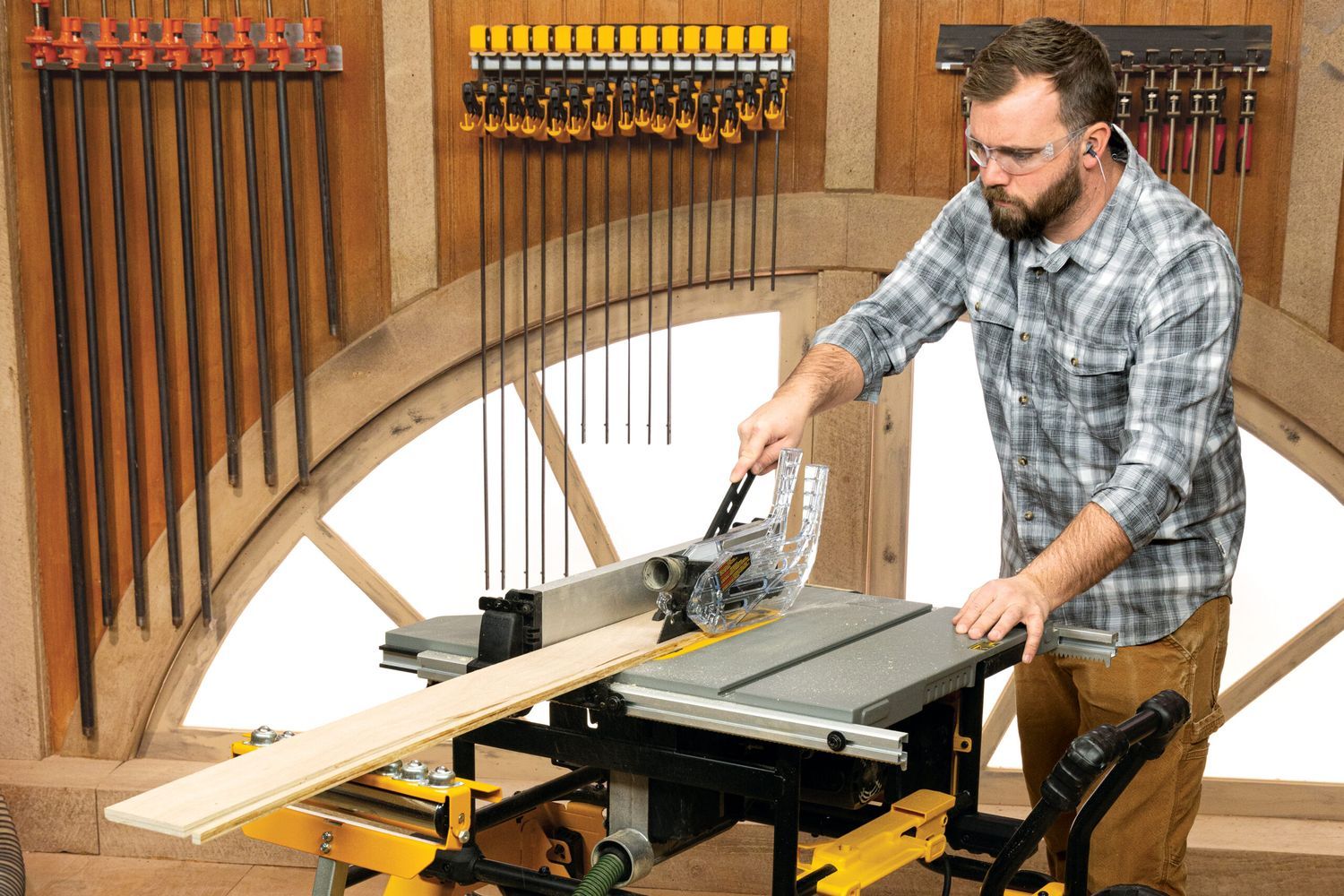 In a well-organized woodshop, a man wearing safety goggles and ear protection expertly operates a compact table saw, showcasing how to choose and use the tool effectively as he cuts a long wooden plank amidst an array of tools hanging on the walls.
