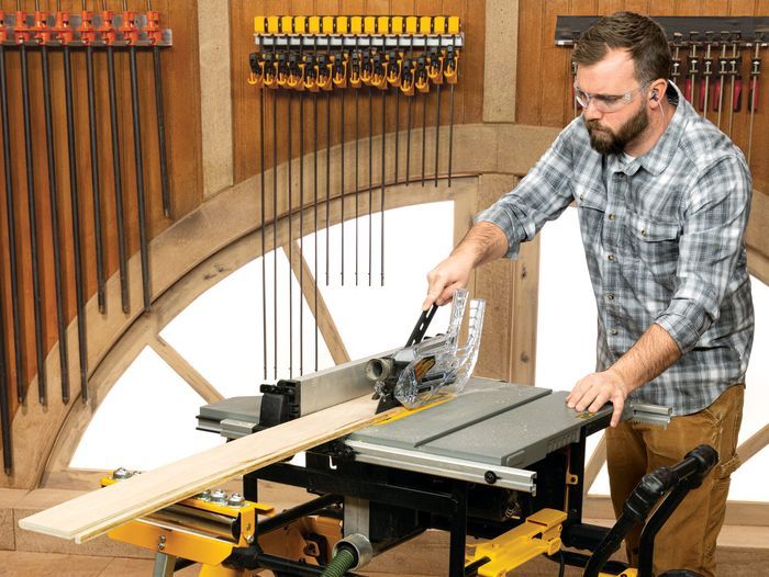 In a well-organized woodshop, a man wearing safety goggles and ear protection expertly operates a compact table saw, showcasing how to choose and use the tool effectively as he cuts a long wooden plank amidst an array of tools hanging on the walls.