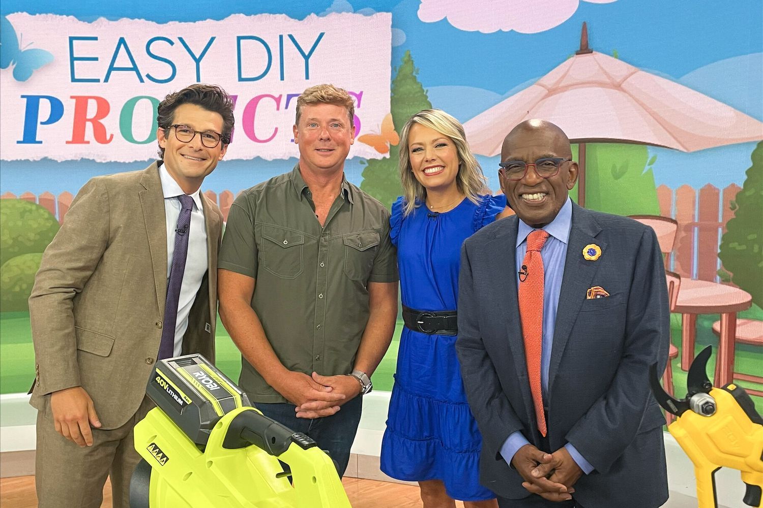 Four people stand together on a TV set labeled "Easy DIY Projects," a segment on the Today Show. Kevin O'Connor, the host of "The Old House" shares his DIY tips. In front of them is a green leaf blower and another tool, while the backdrop features a garden scene with a fence and umbrella.