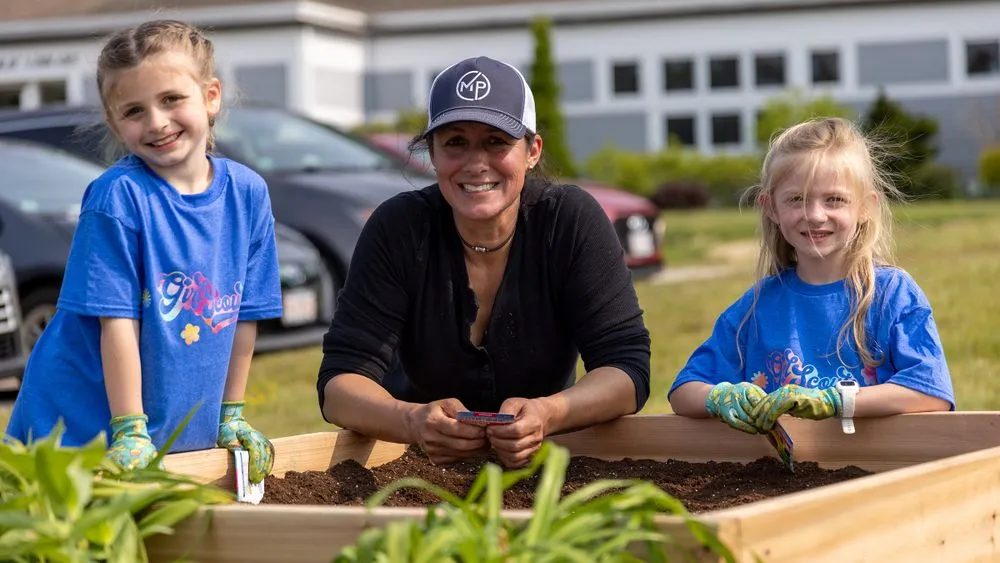 A woman and two young girls from the local Girl Scouts troop are smiling while gardening in a raised wooden bed. The girls are wearing blue shirts and gloves, while the woman sports a black shirt and cap. Behind them, a school building and parked cars complete the community project scene.