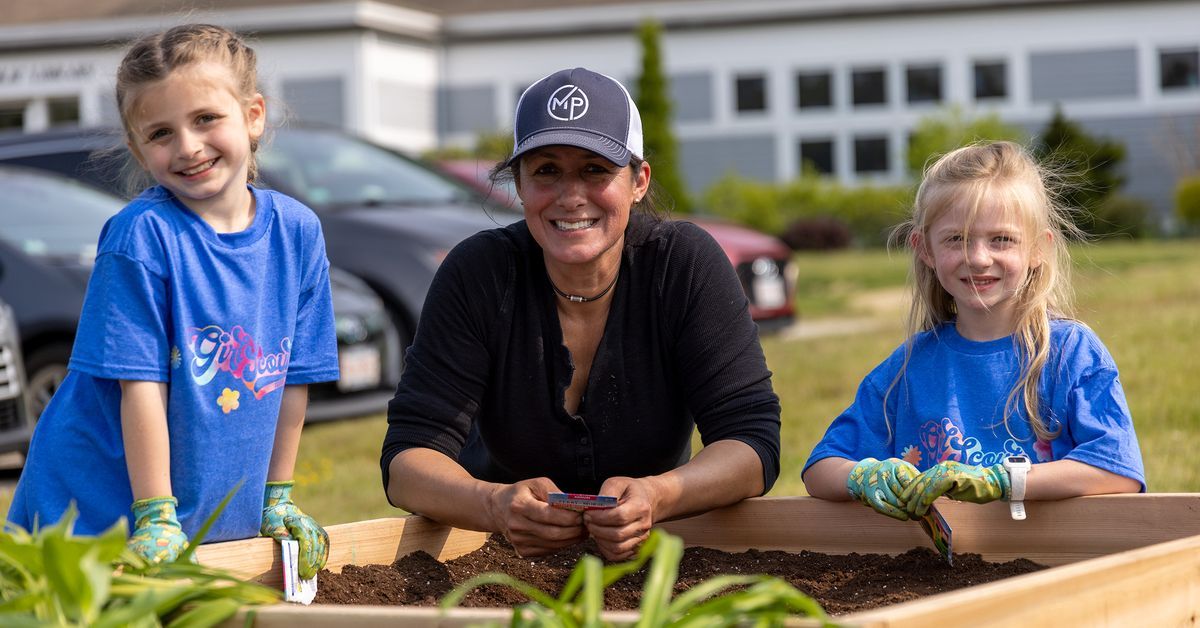 A woman and two young girls from the local Girl Scouts troop are smiling while gardening in a raised wooden bed. The girls are wearing blue shirts and gloves, while the woman sports a black shirt and cap. Behind them, a school building and parked cars complete the community project scene.