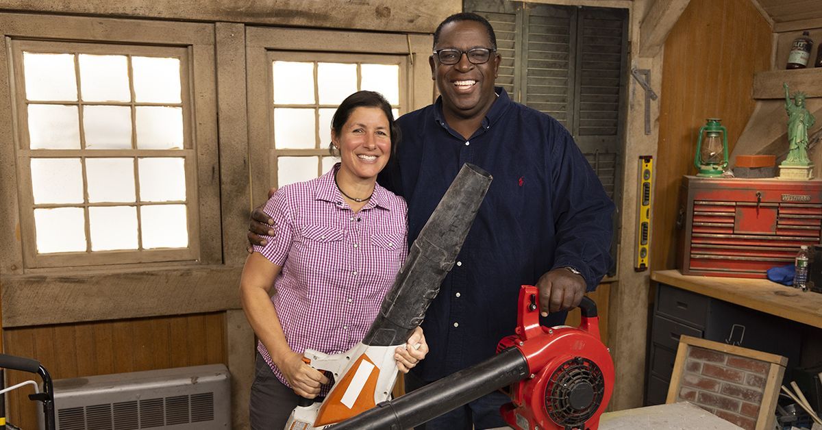 Two people smiling in a workshop. One holds a leaf blower while the other drapes an arm around them.