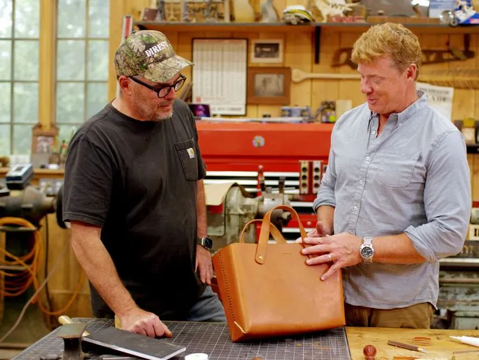 Two men stand in a workshop. One wears a cap and T-shirt; the other wears a light blue shirt. Tools and machinery are visible in the background.