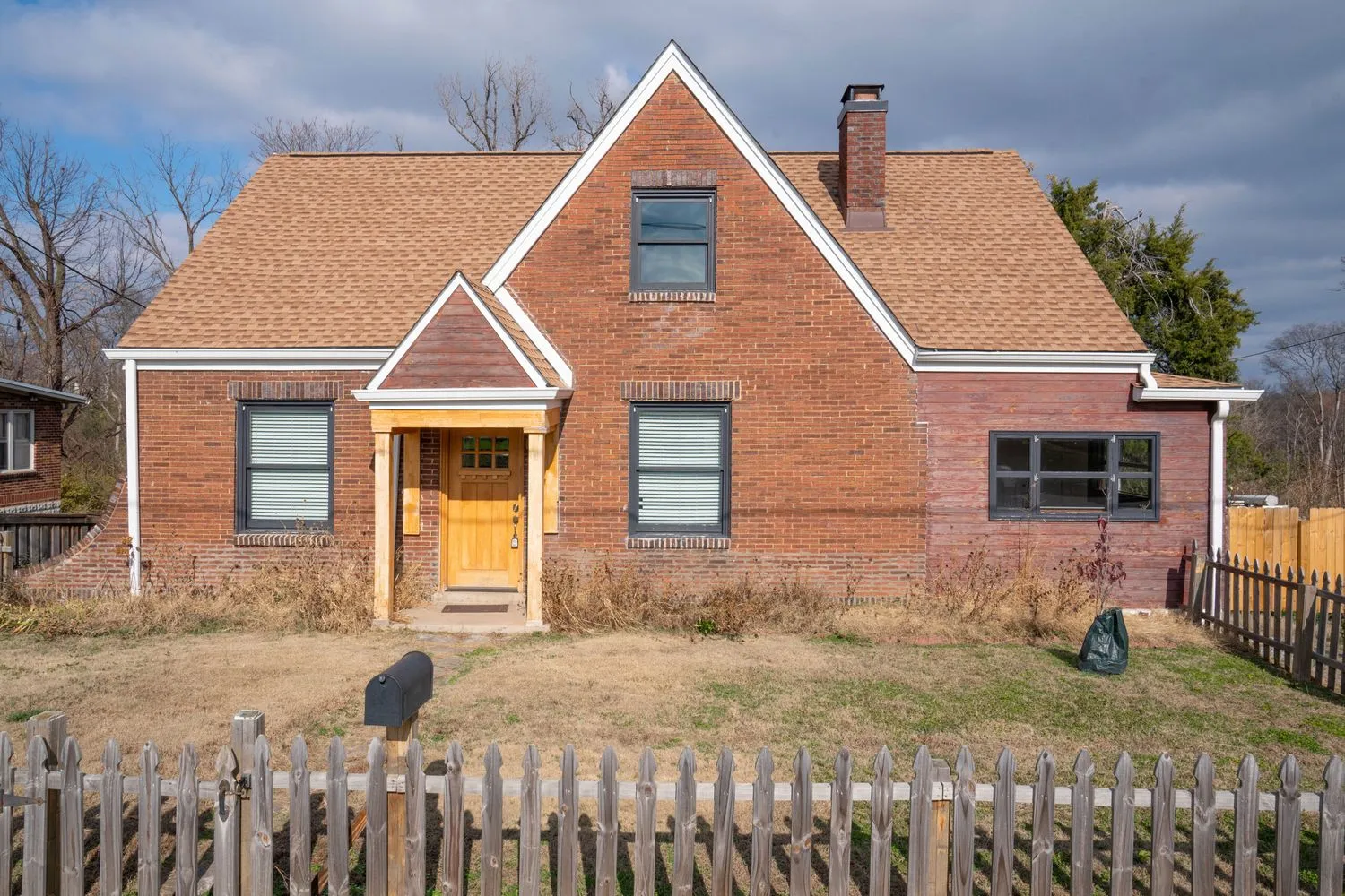 Get a first look at the Nashville Brick Cottage Project House, featuring a red brick facade and steeply pitched brown roof. Its wooden front door is flanked by windows with shutters, and a charming picket fence surrounds the yard with dry grass.