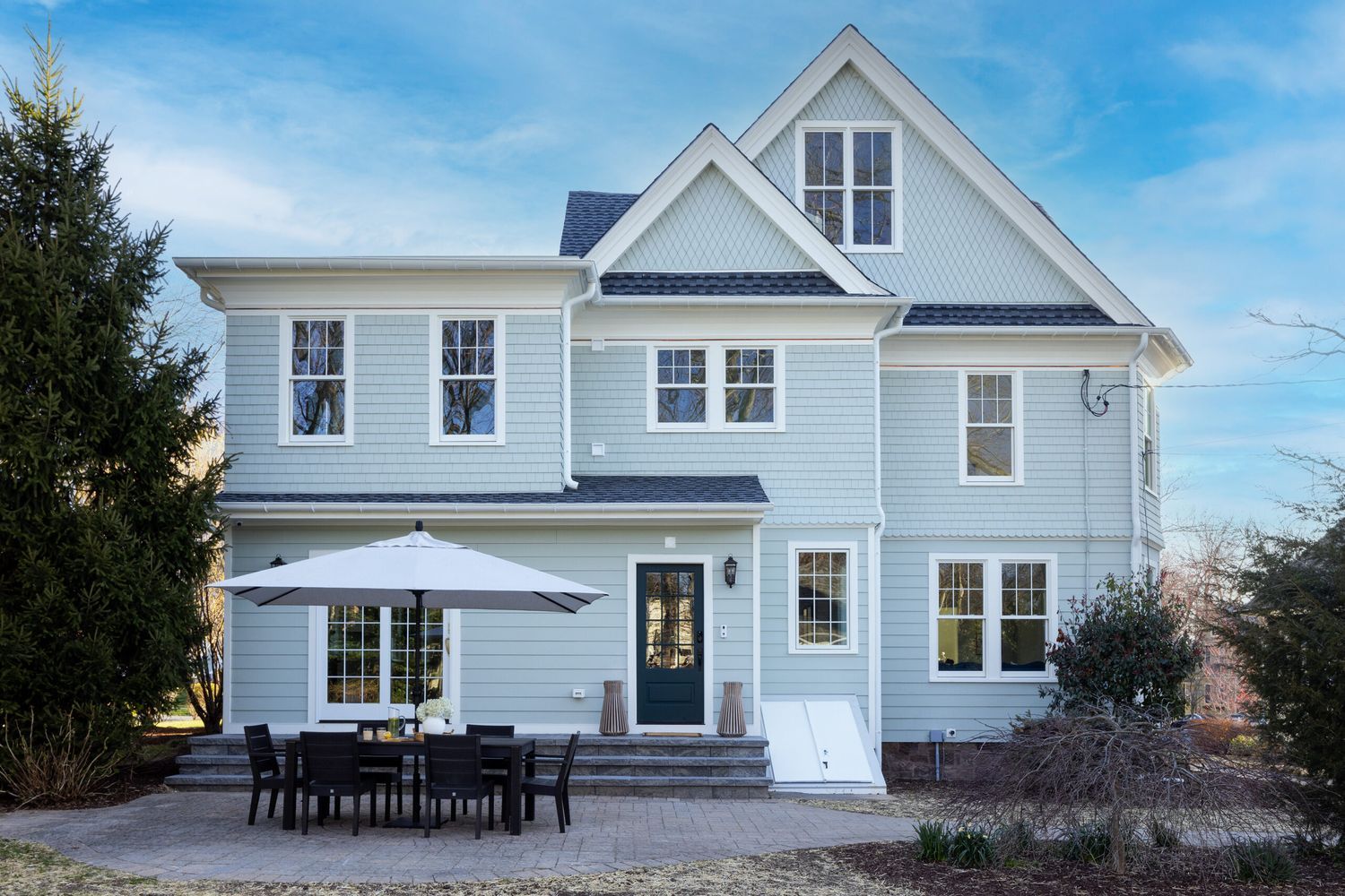 A two-story light gray house with Replacement Windows Done Right features multiple windows, a dark front door, and a sloped roof. A patio with a dining set sits in the backyard under a large white umbrella. Green trees and bushes surround the area under a blue sky.