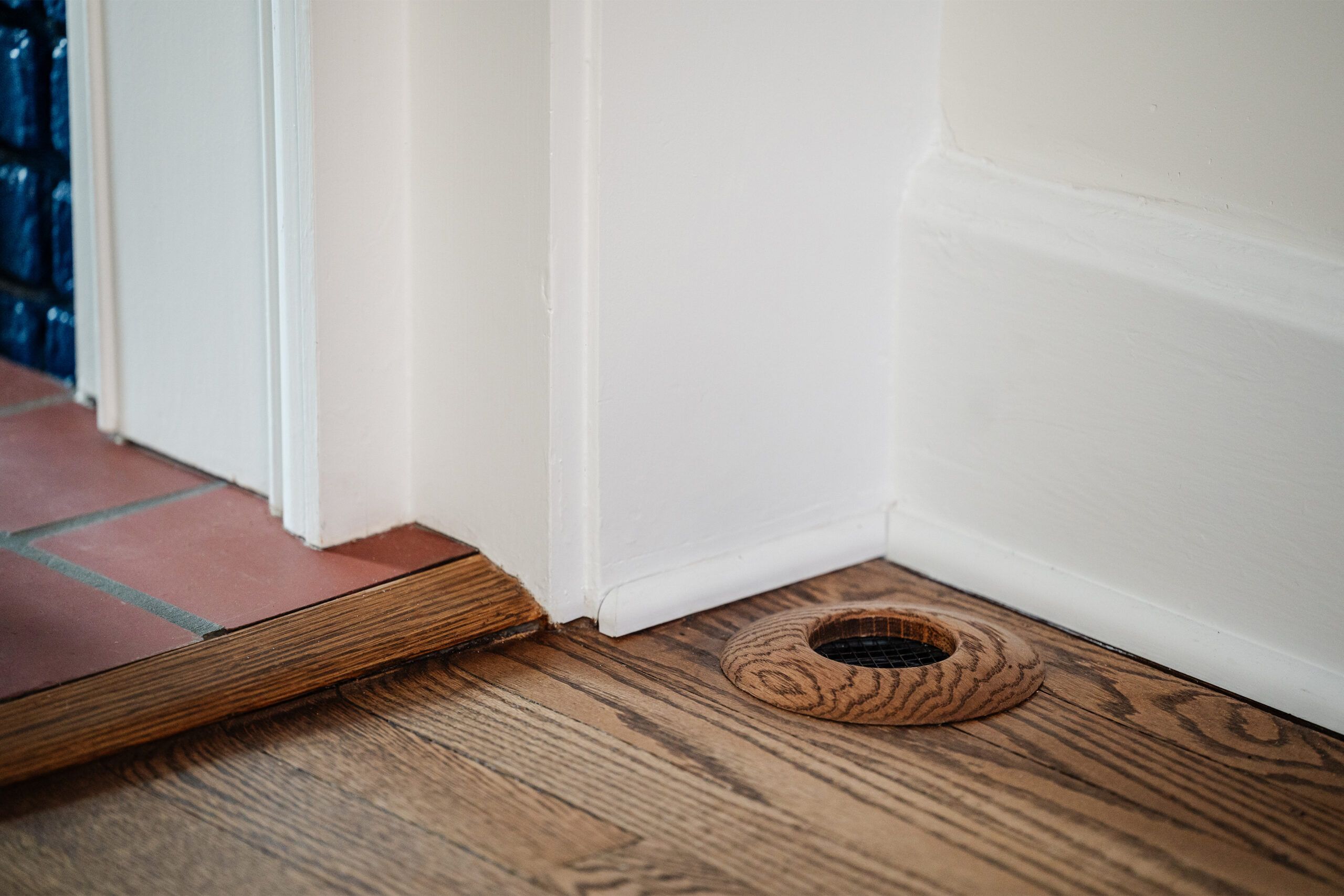 A wooden floor vent, which subtly carries the house smells, is installed on an oak floor near a white wall corner. To the left, there's a transition to terracotta floor tiles.
