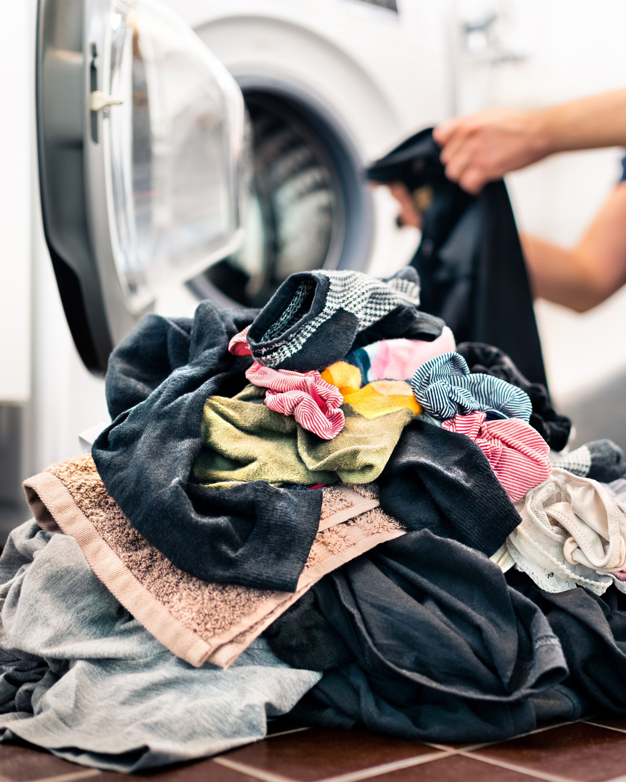 A pile of colorful laundry lies on the floor in front of an open washing machine. A person holds a black garment, ready to load it, as the familiar house smells of fresh detergent fill the air, completing the scene of a typical laundry day at home.