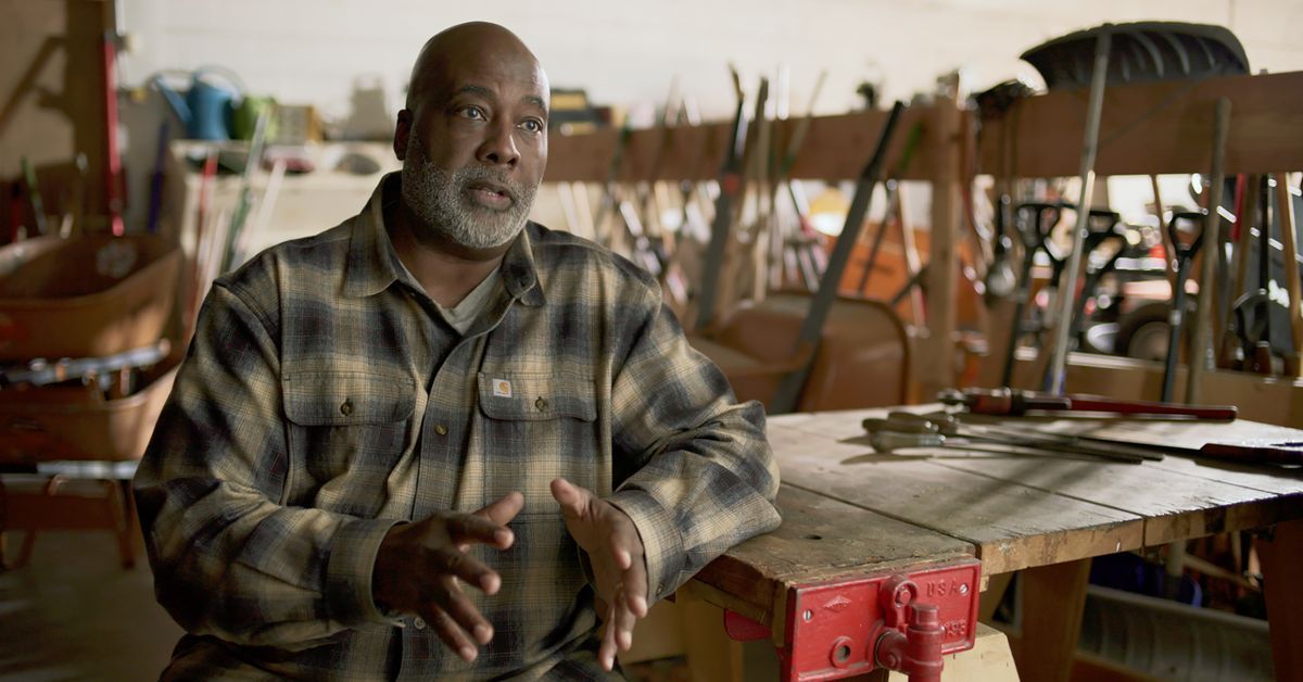 A man with a gray beard, wearing a plaid shirt, sits in a workshop filled with tools and gardening equipment. He gestures with one hand while speaking. A workbench with various tools is in front of him.